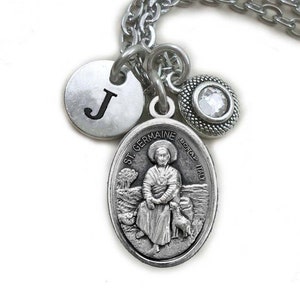 May include: A silver-coloured necklace featuring a pendant with the image of St. Germaine, a round charm with the letter "J", and a decorative charm with a clear crystal. The chain is a linked design.