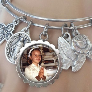 Op de afbeelding: Zilveren armband met verschillende bedels, waaronder een kruis, een religieuze medaille, een roos, een duif en een fotobedel van een jong meisje in een witte jurk. De armband is om de pols van een mannequin.