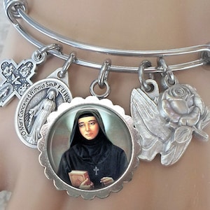 Peut inclure: Un bracelet extensible en argent avec des breloques, dont une croix, une rose, une colombe et un breloque rond avec une image d'une femme en habit noir. Le breloque rond porte le texte "O Mary Conceived Without Sin, P. F. U. W."