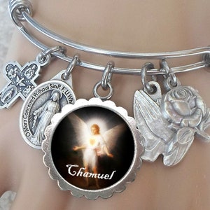 Puede incluir: Un brazalete de plata con varios encantos, incluyendo una cruz, una rosa, una paloma y un encanto redondo con una imagen de un ángel y el texto "Chamuel".
