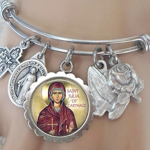 Puede incluir: Pulsera de plata con varios charms, incluyendo una cruz, una rosa, una paloma y un charm redondo con una imagen de Santa Julia de Cartago. La pulsera es ajustable.
