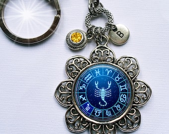 Puede incluir: Un llavero plateado con un dije zodiacal azul y plateado que presenta un escorpión y los símbolos astrológicos de cada signo. El dije está unido a un anillo plateado con un pequeño strass amarillo.