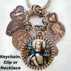 Puede incluir: Un brazalete de charms de bronce con una imagen de vitral de Santa Adela, un charm de corazón, un charm de cruz y un charm con las palabras "Oh María concebida sin pecado ruega por nosotros".