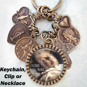 May include: A bronze-toned keychain with a variety of charms, including a heart, a cross, and a round charm with a picture of a woman. The round charm has the text "BLESSED ANNA MARIA TIAGI" on it. The keychain can also be used as a clip or necklace.