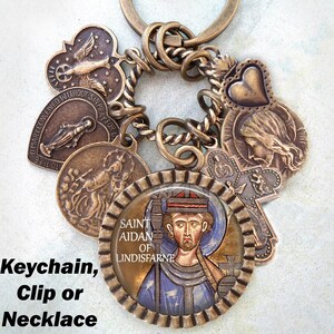 Può includere: Un portachiavi dorato con una varietà di ciondoli, tra cui un cuore, una croce e un ciondolo rotondo con un'immagine dipinta di San Aidan di Lindisfarne. Il testo "SAINT AIDAN OF LINDISFARNE" è visibile sul ciondolo rotondo. Il portachiavi può essere utilizzato come collana o clip.