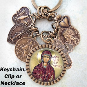 Puede incluir: Un llavero de bronce con una variedad de dijes, incluyendo un corazón, una cruz y un dije redondo con una imagen de Santa Julia de Cartago. Los dijes están unidos a una cadena de bronce retorcida.