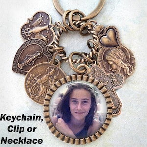 May include: A bronze-toned keychain featuring a selection of charms, including a heart, a cross, and a round photo frame. The photo frame displays a picture of a young person with brown hair. The keychain is labelled "Keychain, Clip or Necklace".