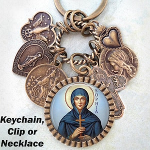 Può includere: Un portachiavi in bronzo con una varietà di ciondoli, tra cui un cuore, una croce e un ciondolo rotondo con l'immagine di una donna in una veste blu che tiene una croce. Il ciondolo rotondo ha la scritta "SOPHIA" su di esso.