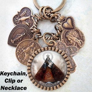 May include: A bronze-toned keychain with a variety of charms, including a heart, a cross, and a round charm with a picture of the Virgin Mary. The keychain is attached to a twisted metal ring. The text "O MARY CONCEIVED WITHOUT SIN PRAY FOR US" and "AS DE LA CARIDAD DEL CORRAL" are visible on the charms.