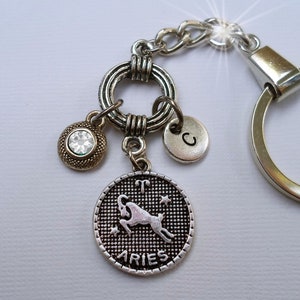 May include: Silver keychain with a round charm featuring the zodiac symbol for Aries, a ram, with the word "Aries" below it. The charm also has a small silver charm with the letter "C" and a round charm with a clear rhinestone.