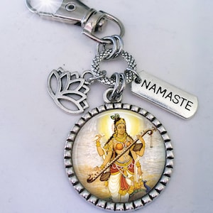May include: Silver keychain with a round charm featuring a colorful image of a Hindu goddess playing a stringed instrument. The charm is surrounded by a silver border and has a silver lotus flower charm and a silver tag with the word "NAMASTE" on it.