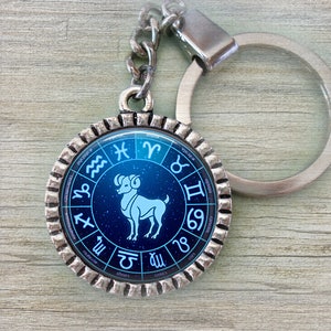 May include: Silver keychain with a blue and silver circular charm featuring the zodiac symbol for Aries. The charm has a blue background with a white Aries symbol and a silver ring around the edge. The keychain is attached to a silver ring.