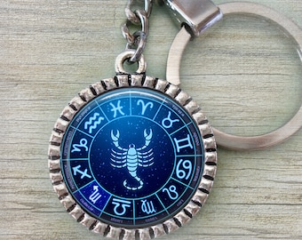 Puede incluir: Un llavero plateado con un colgante redondo que representa el signo zodiacal de Escorpio. El colgante tiene un fondo azul con un escorpión blanco y símbolos del zodiaco. El llavero incluye una cadena y un anillo.