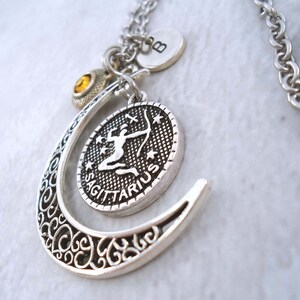 Puede incluir: Collar con colgante plateado del signo zodiacal Sagitario. El colgante presenta un dise&ntilde;o de arquero detallado dentro de un marco ovalado, acentuado por una forma de media luna de filigrana. Los dijes adicionales incluyen un disco inicial y una gema amarilla.