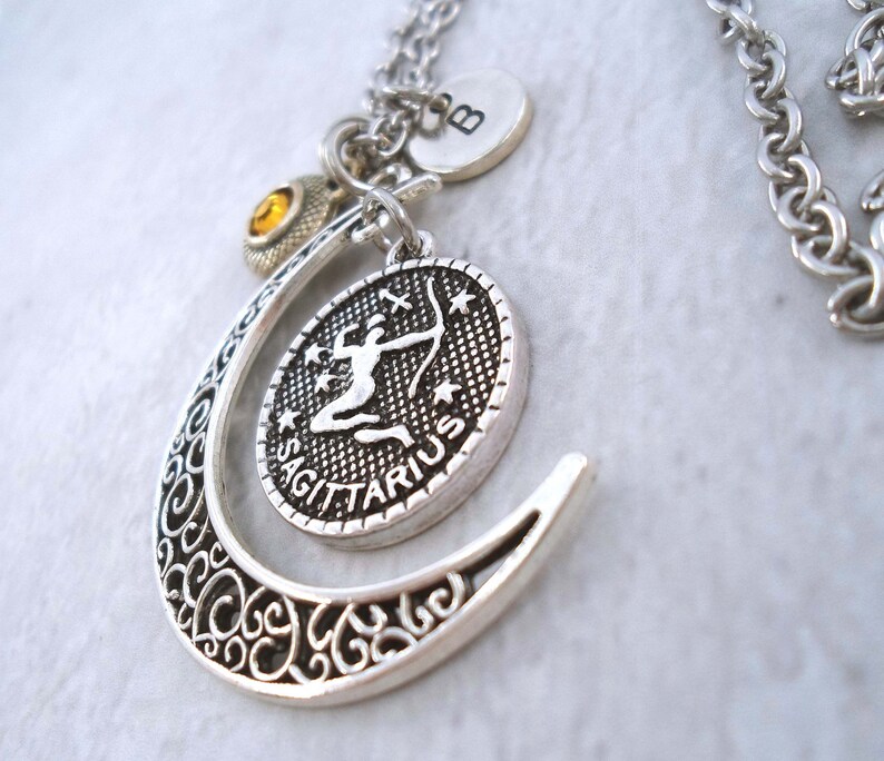 Puede incluir: Collar con colgante plateado del signo zodiacal Sagitario. El colgante presenta un dise&ntilde;o de arquero detallado dentro de un marco ovalado, acentuado por una forma de media luna de filigrana. Los dijes adicionales incluyen un disco inicial y una gema amarilla.