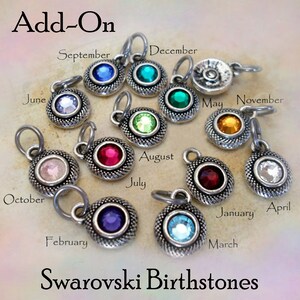 Op de afbeelding: Twaalf zilveren bedels, elk met een kristallen geboortesteen en gelabeld met een maand van het jaar. De bedels zijn in een cirkel op een lichtbruin oppervlak gerangschikt. De tekst "Swarovski Birthstones" staat onderaan de afbeelding.