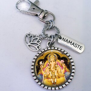 Op de afbeelding: Zilveren sleutelhanger met een ronde hanger met een kleurrijk beeld van de hindoe-god Ganesha. De hanger is omgeven door een zilveren rand met een decoratief patroon. De sleutelhanger bevat ook een zilveren lotusbloemhanger en een zilveren label met het woord "NAMASTE" erop gegraveerd.