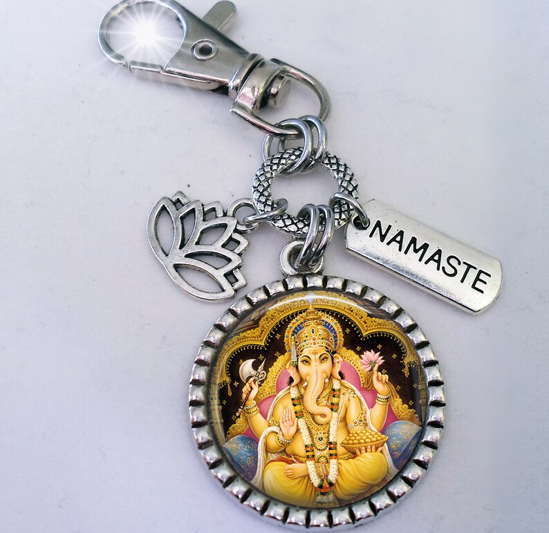 Op de afbeelding: Zilveren sleutelhanger met een ronde hanger met een kleurrijk beeld van de hindoe-god Ganesha. De hanger is omgeven door een zilveren rand met een decoratief patroon. De sleutelhanger bevat ook een zilveren lotusbloemhanger en een zilveren label met het woord "NAMASTE" erop gegraveerd.
