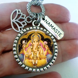 Op de afbeelding: Zilveren bedel met een ronde afbeelding van de hindoe-god Ganesha, omringd door een decoratieve rand. De bedel is bevestigd aan een zilveren ketting met een lotusbloembedel en een tag met de tekst "NAMASTE".