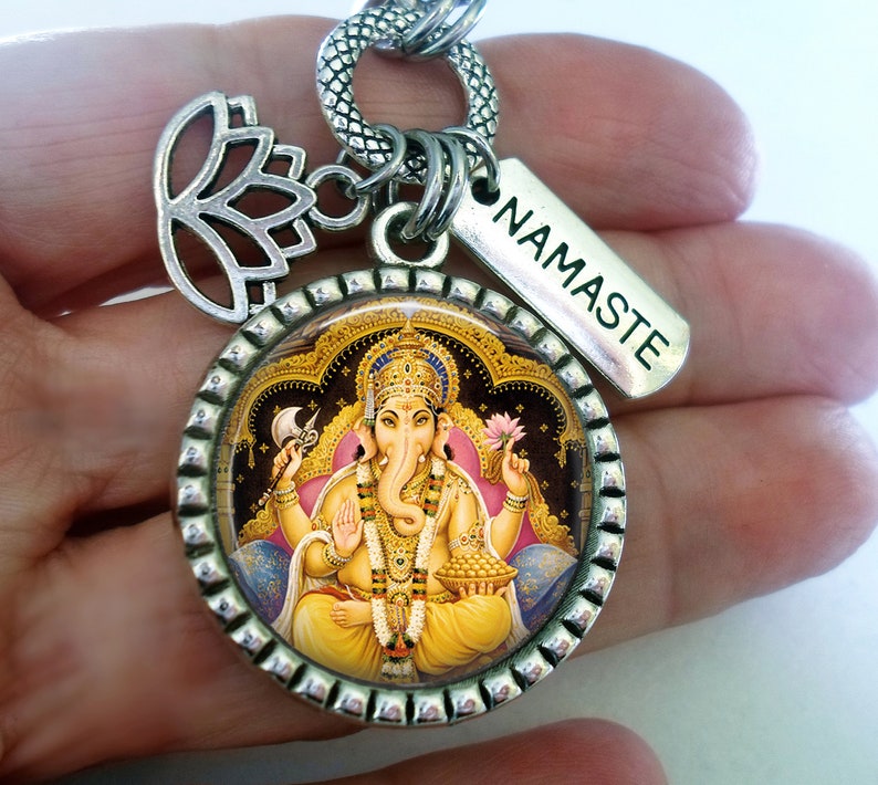 Op de afbeelding: Zilveren bedel met een ronde afbeelding van de hindoe-god Ganesha, omringd door een decoratieve rand. De bedel is bevestigd aan een zilveren ketting met een lotusbloembedel en een tag met de tekst "NAMASTE".