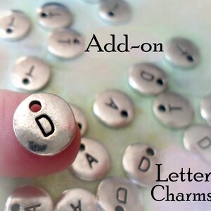 Op de afbeelding: Zilveren letterbedels met een gaatje om aan sieraden of andere knutselwerkjes te bevestigen. De bedels zijn rond met een letter in het midden. De letters die te zien zijn, zijn A en D. De tekst "Add-on Letter Charms" is zichtbaar in de afbeelding.