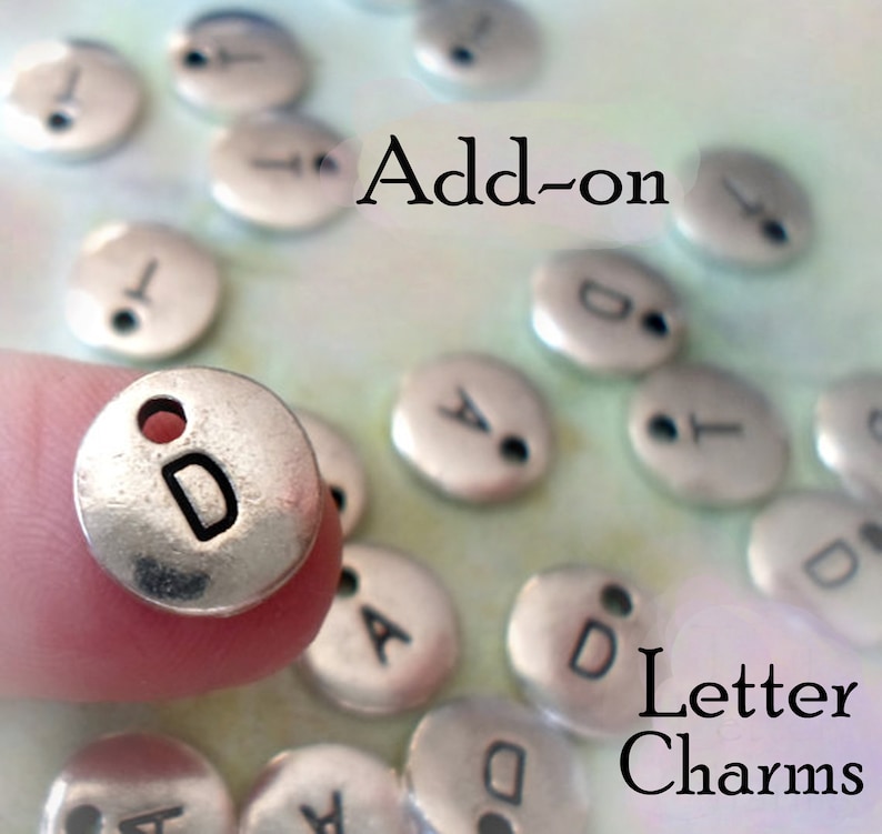 Op de afbeelding: Zilveren letterbedels met een gaatje om aan sieraden of andere knutselwerkjes te bevestigen. De bedels zijn rond met een letter in het midden. De letters die te zien zijn, zijn A en D. De tekst "Add-on Letter Charms" is zichtbaar in de afbeelding.