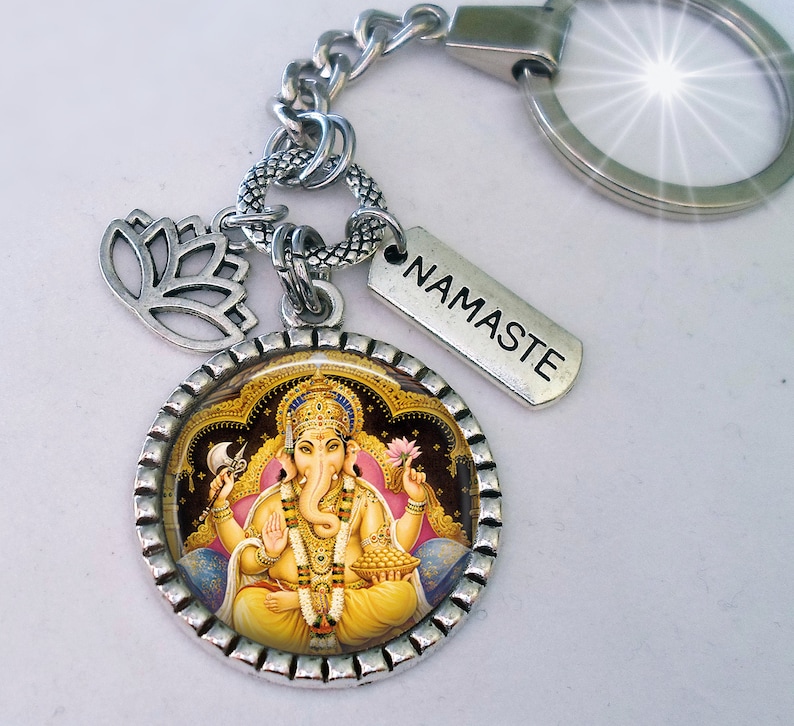 Op de afbeelding: Zilveren sleutelhanger met een ronde hanger met een kleurrijk beeld van de hindoe-god Ganesha. De hanger is omgeven door een zilveren rand met een decoratief patroon. De sleutelhanger bevat ook een zilveren lotusbloemhanger en een zilveren tag met het woord "Namaste" erop gegraveerd.