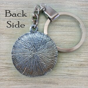 May include: Silver-toned metal keychain with a round, textured charm and a silver-toned metal ring. The back of the charm is visible.