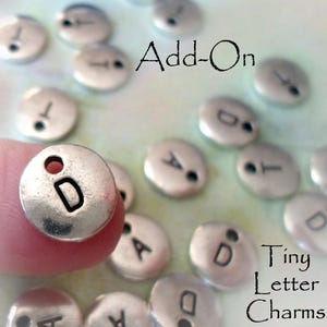 May include: Silver metal letter charms with a hole for stringing. The charms are round and have a slightly textured surface. The letters are in uppercase and are centered on the charms. The charms are labeled "Add-On Tiny Letter Charms".