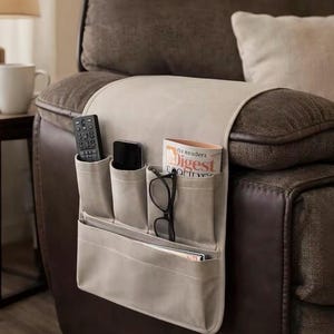 May include: Beige fabric sofa armrest organiser with multiple pockets. The organiser holds a remote control, a smartphone, reading glasses, and a magazine. The armrest is attached to a brown leather recliner.