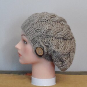 Könnte beinhalten: Eine beige Strickmütze mit Zopfmuster und einem Knopf aus Holz.