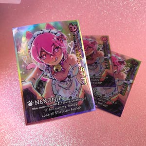 May include: Holographic trading cards featuring a pink-haired anime character in a maid outfit with cat ears. The card has the text "NEKONIP" and "STT 26". The card also has the text "Mm-mm-mmmh! You're smelling a lil' too yummy, buddy. Lose all STA, Gain full HP."