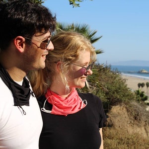 May include: A couple wearing sunglasses and face masks stand side-by-side, looking out at a beach scene with palm trees and the ocean in the distance.