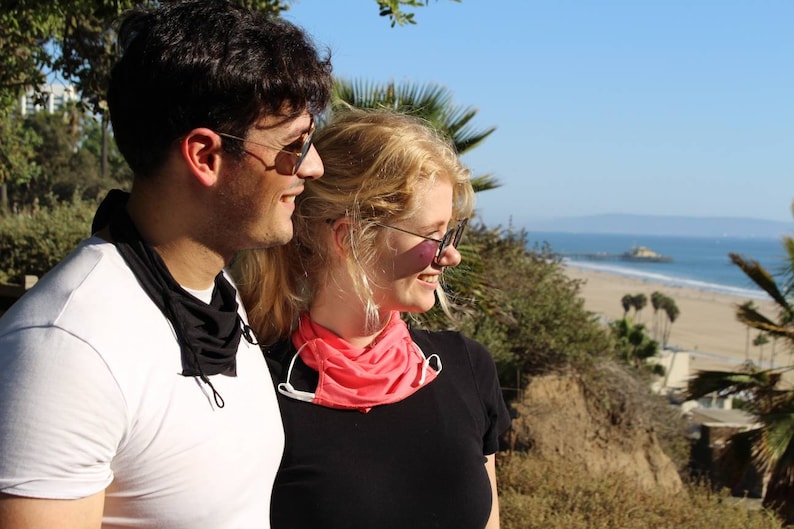 May include: A couple wearing sunglasses and face masks stand side-by-side, looking out at a beach scene with palm trees and the ocean in the distance.