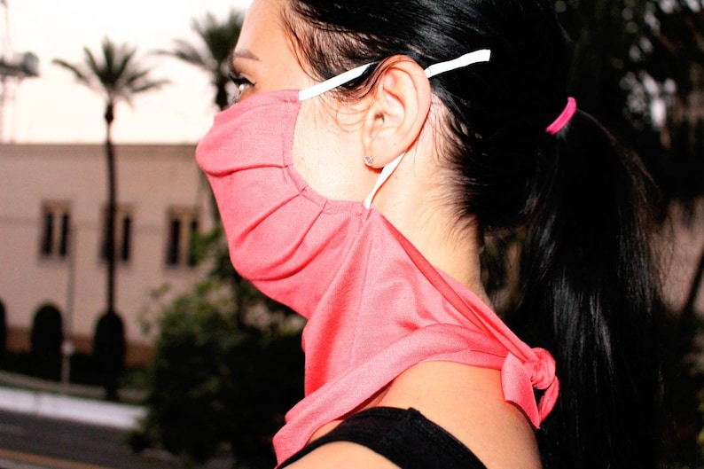 May include: A pink face mask with white straps, worn around the neck and tied at the back. The mask is made of a soft, stretchy fabric.