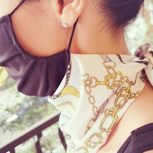 May include: A white scarf with a gold chain pattern and a black face mask. The scarf is tied around the neck and the mask is covering the mouth and nose.