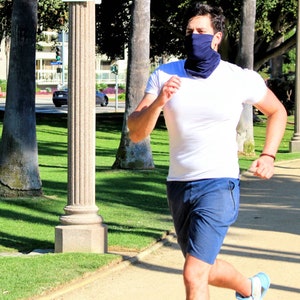 May include: A person wearing a white t-shirt and blue shorts is running outdoors. They are wearing a blue face covering.