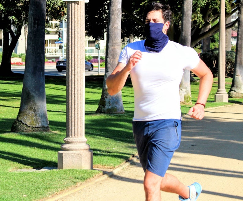 May include: A person wearing a white t-shirt and blue shorts is running outdoors. They are wearing a blue face covering.
