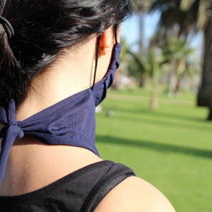 May include: A person wearing a dark blue face mask tied around their neck. The mask is made of a soft, stretchy fabric. The person is standing in a grassy area with palm trees in the background.