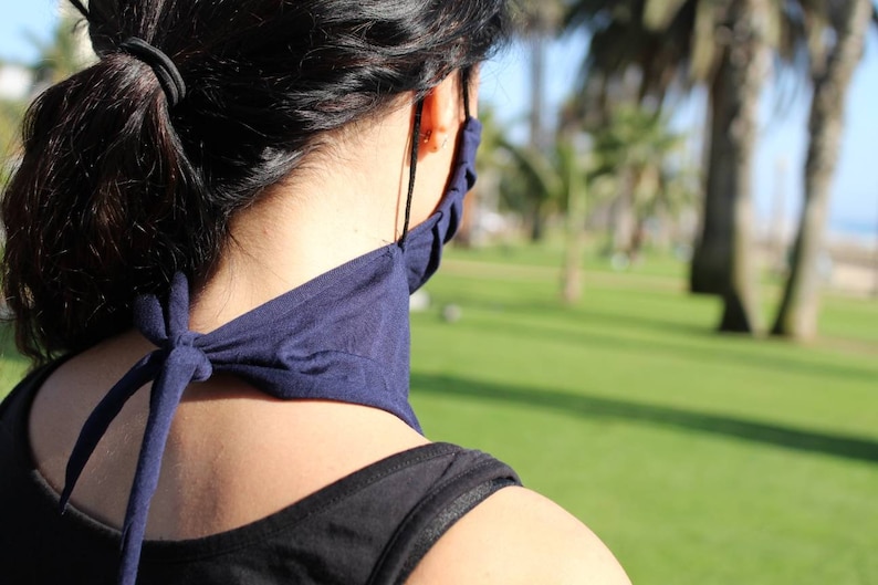 May include: A person wearing a dark blue face mask tied around their neck. The mask is made of a soft, stretchy fabric. The person is standing in a grassy area with palm trees in the background.