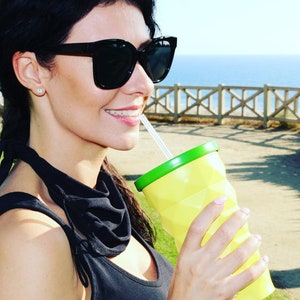 May include: A woman wearing black sunglasses and a black top with a green scarf is drinking from a yellow and green reusable cup with a straw.