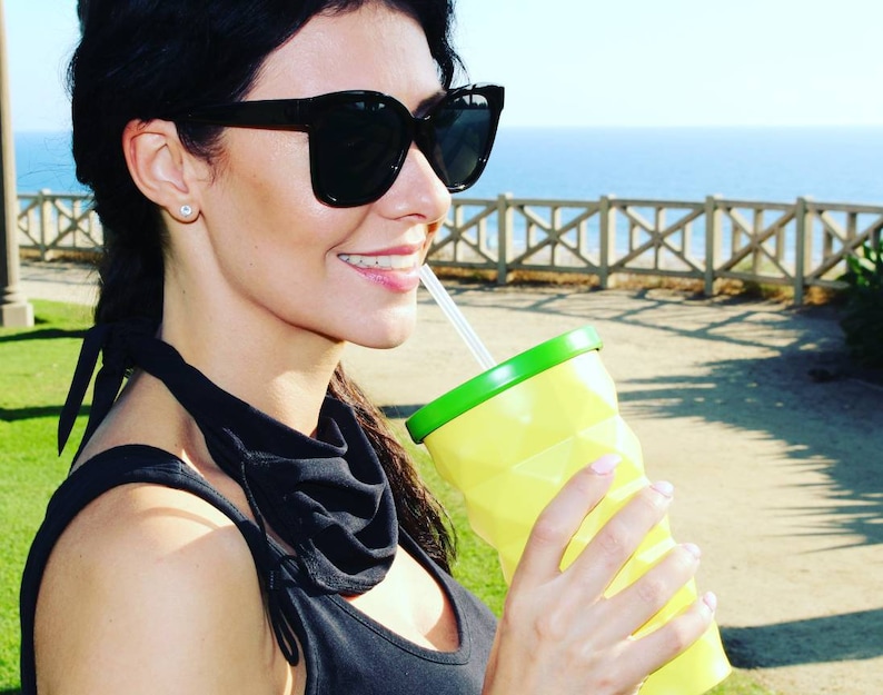 May include: A woman wearing black sunglasses and a black top with a green scarf is drinking from a yellow and green reusable cup with a straw.