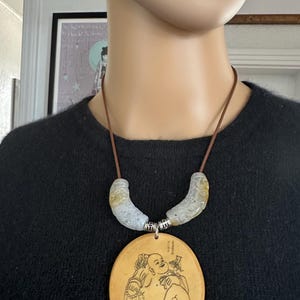 Puede incluir: Collar con un colgante grande y redondo que representa a un Buda sonriente. El colgante está suspendido de un cordón marrón, con dos piezas de jade curvadas y talladas. Este collar único es una joya distintiva.