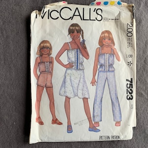 May include: Vintage McCall's sewing pattern featuring illustrations of three outfits: a romper, a dress, and pants with a matching top. The pattern cover is white with the McCall's logo and the price $2.00.