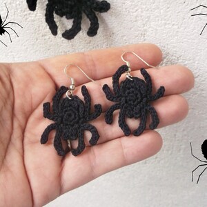 May include: A pair of black crocheted spider earrings hanging from silver hooks. The earrings are held in a hand against a white background.