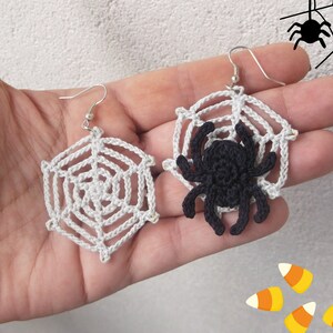 May include: A pair of crocheted spider web earrings with a black crocheted spider on one earring. The earrings are held in a hand against a white background.