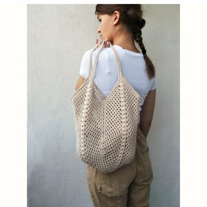 May include: A beige crocheted tote bag with a wide, open top and thick straps. The bag is being worn over the shoulder of a person wearing a white shirt and brown trousers.