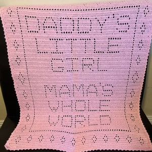 K&ouml;nnte beinhalten: Eine rosa geh&auml;kelte Babydecke mit den Worten "Mama's Girl" in schwarzem Garn.
