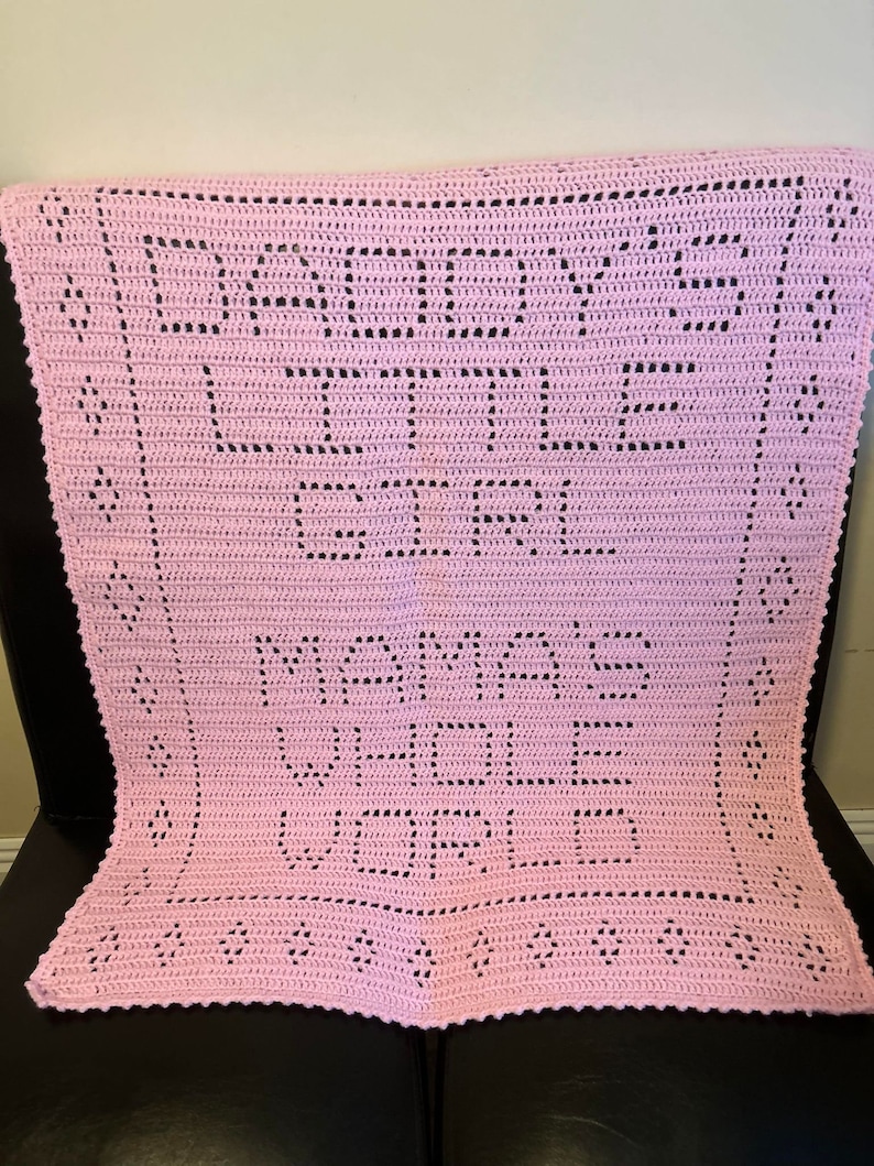K&ouml;nnte beinhalten: Eine rosa geh&auml;kelte Babydecke mit den Worten "Mama's Girl" in schwarzem Garn.