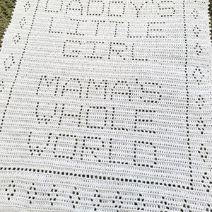 K&ouml;nnte beinhalten: Wei&szlig;e geh&auml;kelte Babydecke mit den Worten "Baby's First Year" und "Mommy's Little Angel" in einer dekorativen Schrift.